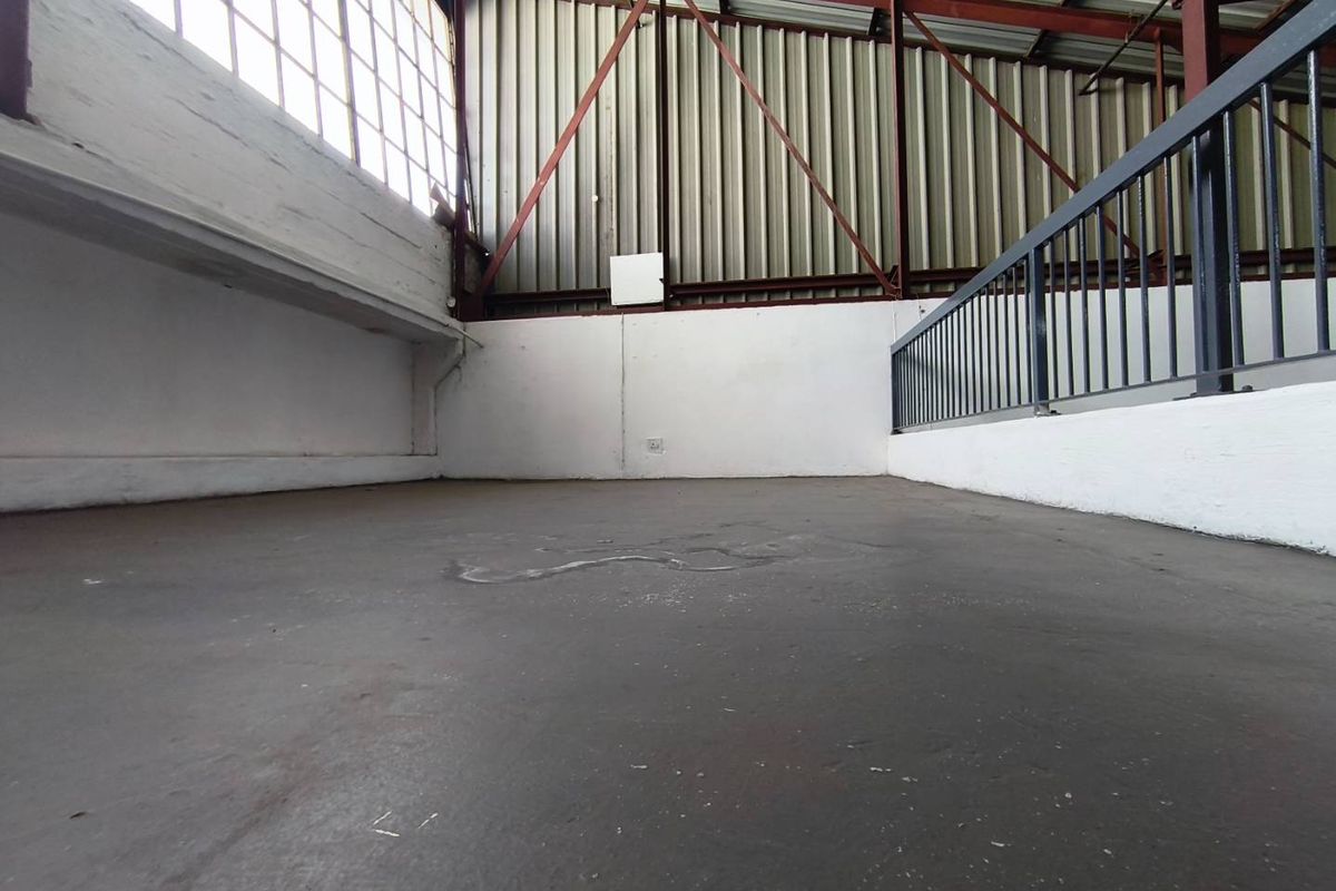 An empty industrial mezzanine level featuring exposed steel trusses, corrugated metal cladding, large steel-framed windows, and a protective metal railing.