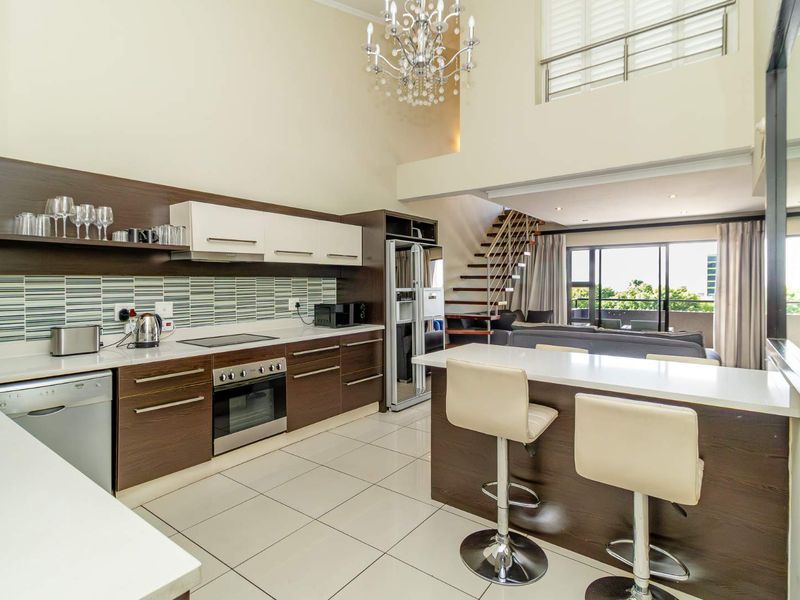 Modern open-plan kitchen and living area featuring sleek cabinetry, stone countertops, a breakfast bar, and a staircase leading to a mezzanine level.