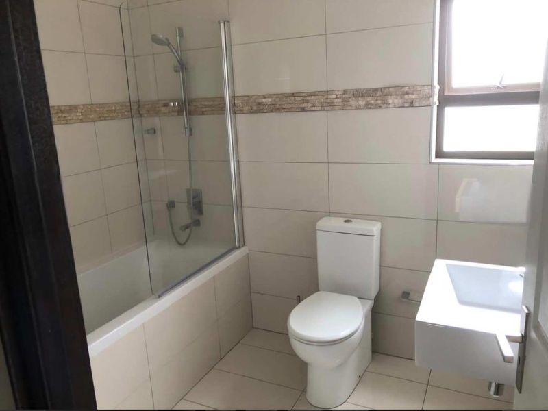 A clean, modern bathroom featuring a bathtub with a glass shower screen, a wall-mounted basin, and a toilet, finished with neutral wall and floor tiles.