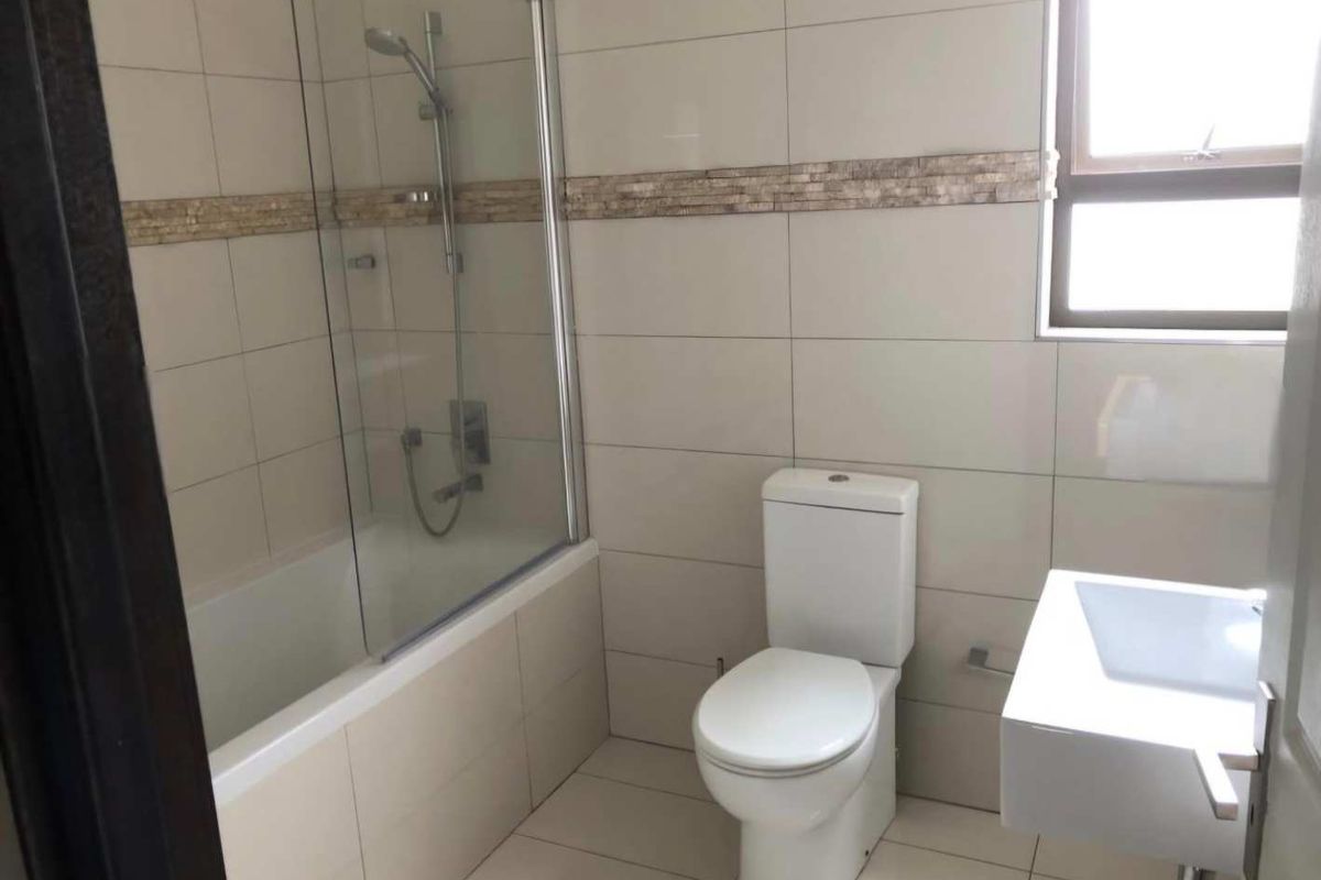 A clean, modern bathroom featuring a bathtub with a glass shower screen, a wall-mounted basin, and a toilet, finished with neutral wall and floor tiles.