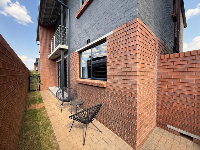 A well-maintained rear exterior view featuring a paved patio area, brick facade, and an upper-level balcony with metal railings.