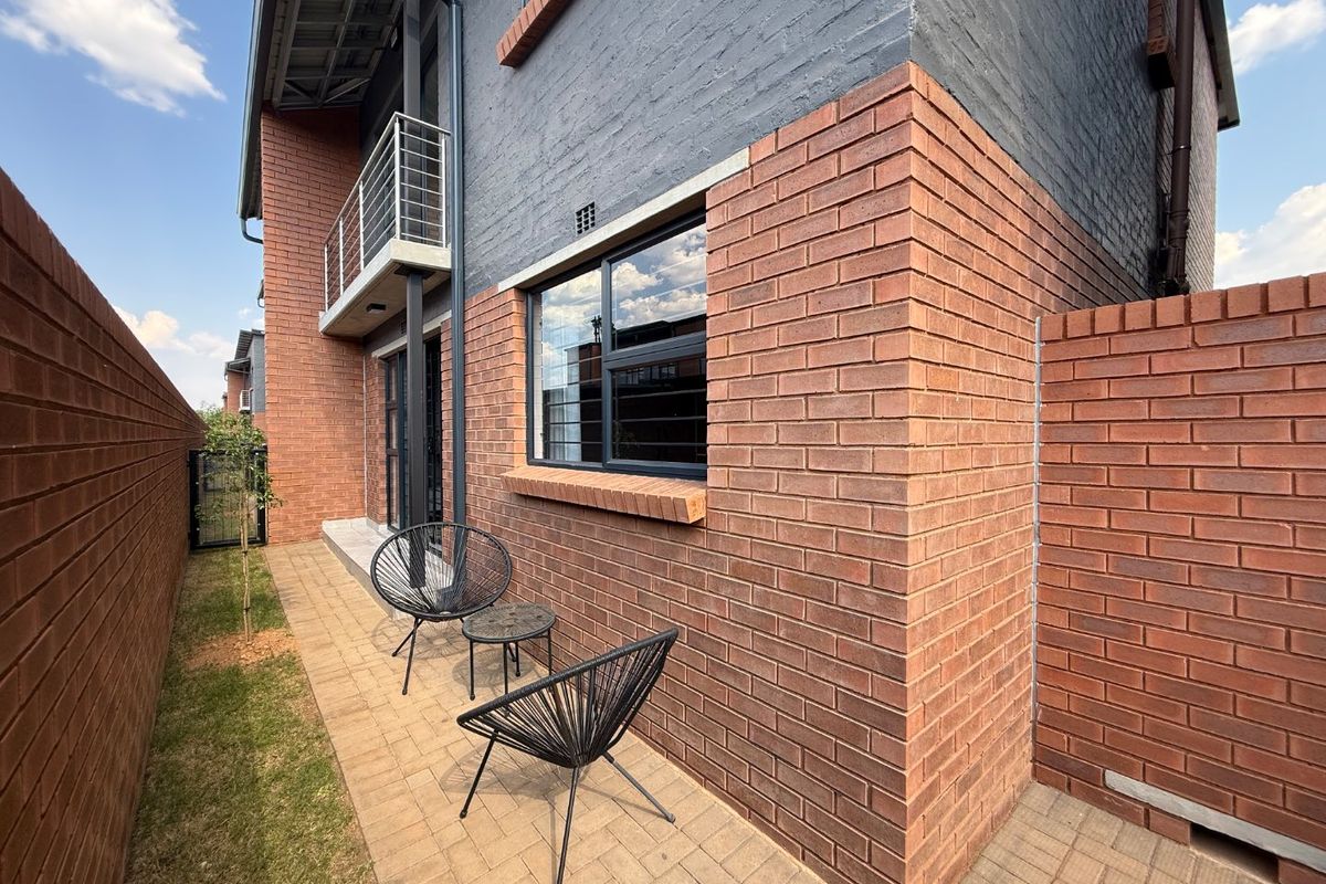 A well-maintained rear exterior view featuring a paved patio area, brick facade, and an upper-level balcony with metal railings.