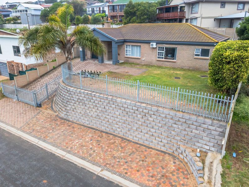 Elevated view of a single-story brick house featuring a paved driveway, secure metal fencing, and a prominent retaining wall in a residential neighborhood.