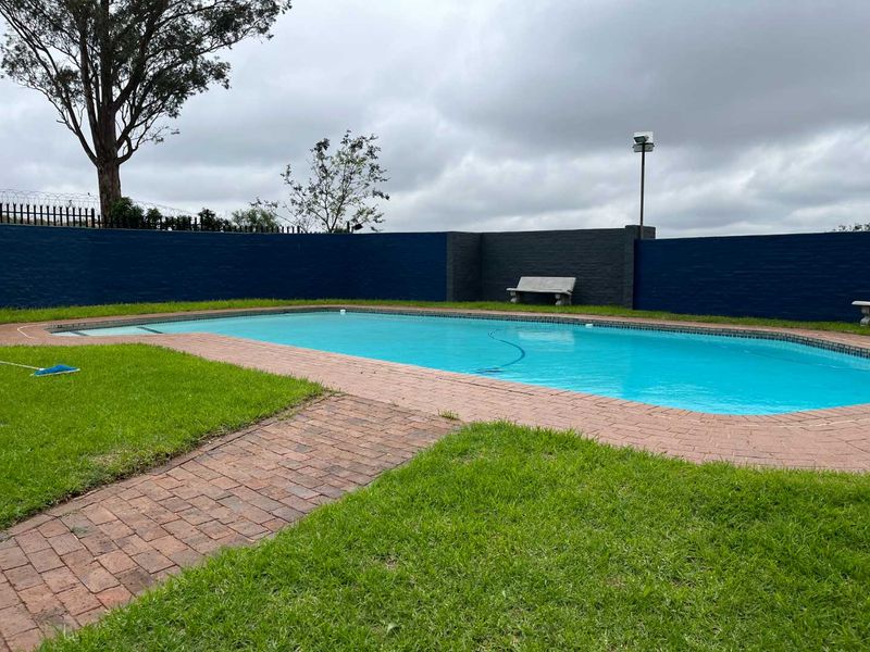 A communal swimming pool area featuring brick paving, a surrounding lawn, and a painted perimeter wall with security fencing.