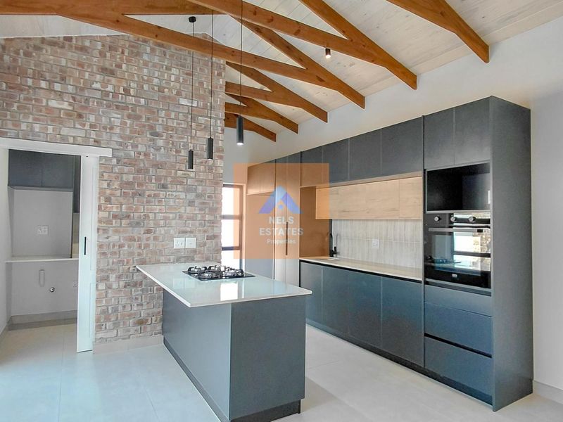 Modern kitchen featuring exposed timber rafters, a brick feature wall, sleek dark cabinetry, stone countertops, and a central island with a built-in gas hob.