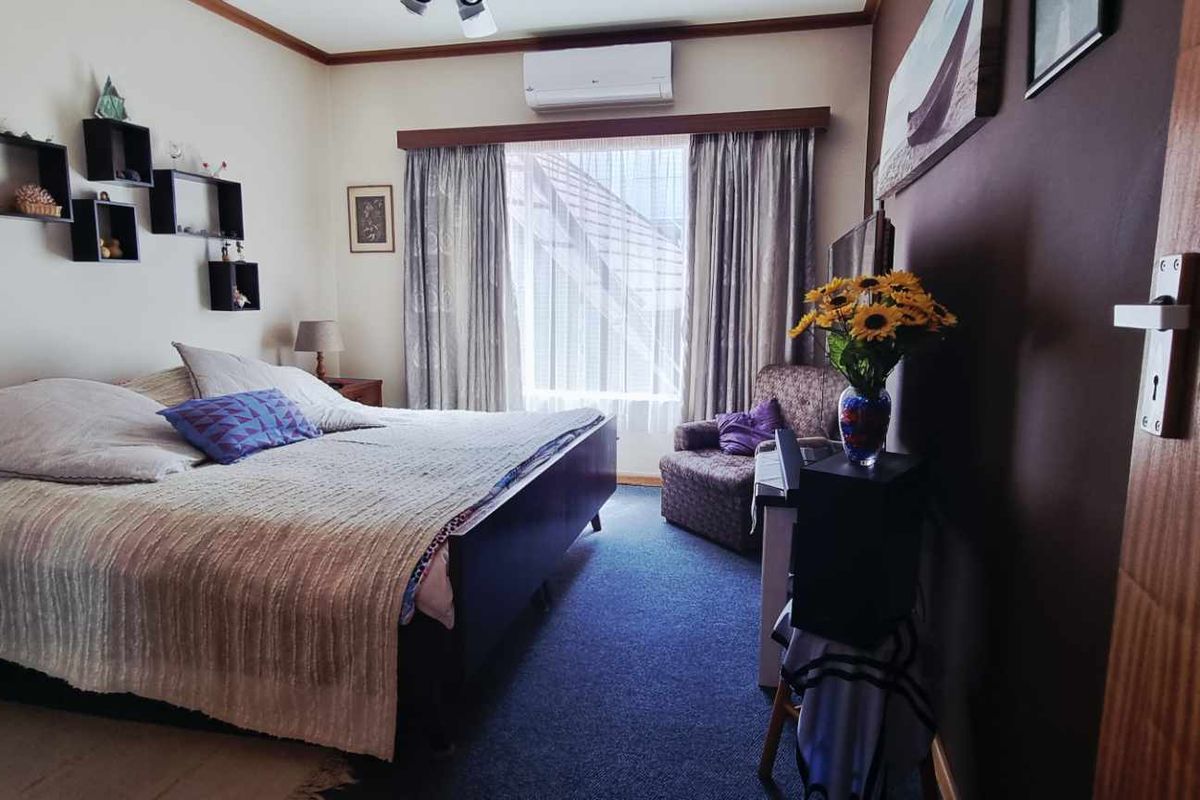 A comfortable bedroom featuring a wall-mounted air conditioning unit, carpeted flooring, and large windows allowing for natural light.