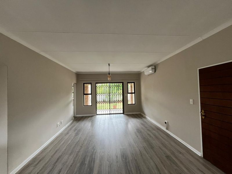 A spacious, empty living room featuring modern laminate flooring, neutral wall tones, an air conditioning unit, and a secure sliding door leading outdoors.