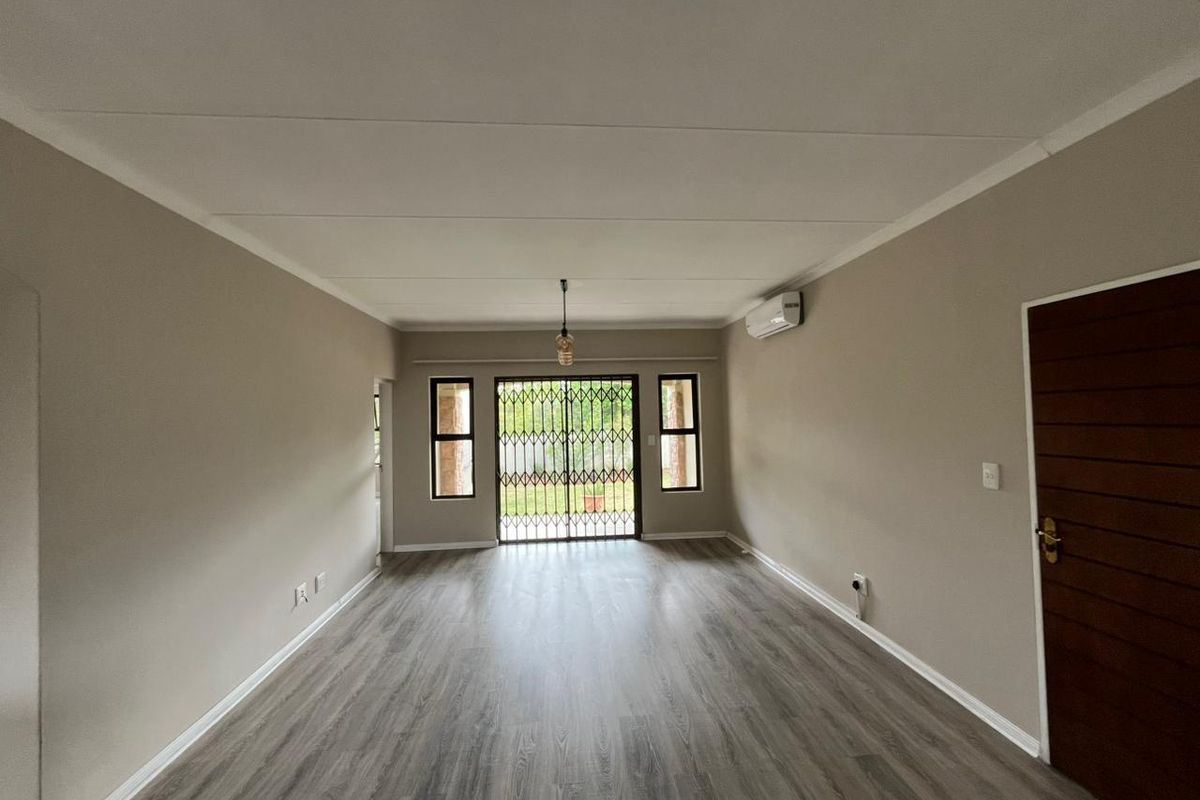 A spacious, empty living room featuring modern laminate flooring, neutral wall tones, an air conditioning unit, and a secure sliding door leading outdoors.