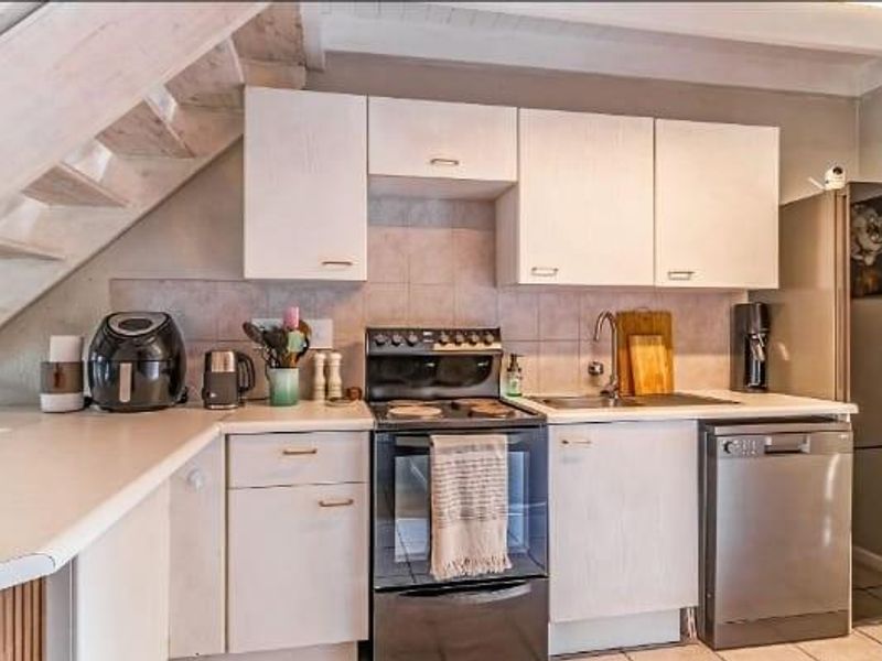 Compact kitchen featuring white cabinetry, tiled backsplash, stainless steel sink, and integrated dishwasher, conveniently positioned beneath a staircase.