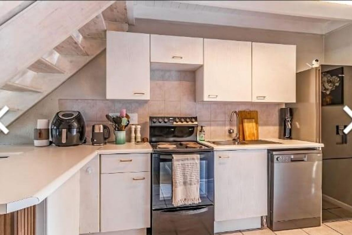 Compact kitchen featuring white cabinetry, tiled backsplash, stainless steel sink, and integrated dishwasher, conveniently positioned beneath a staircase.