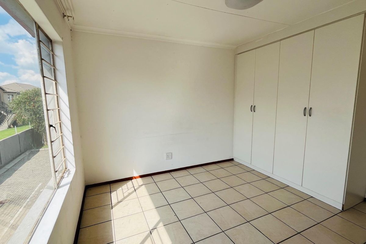 A bright, empty bedroom featuring built-in floor-to-ceiling wardrobes, tiled flooring, and large windows providing natural light and a view of the complex.
