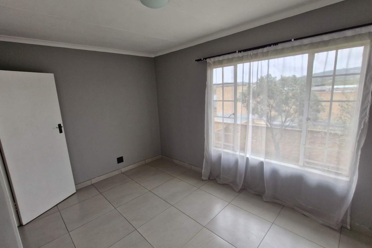 A bright and neutral bedroom featuring large windows with security bars, tiled flooring, and clean grey walls, offering a blank canvas for personalization.