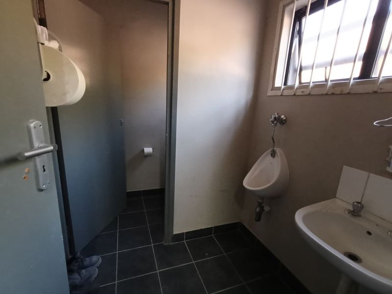 A functional industrial-style bathroom featuring a wall-mounted urinal, pedestal basin, and tiled flooring with security bars on the window.