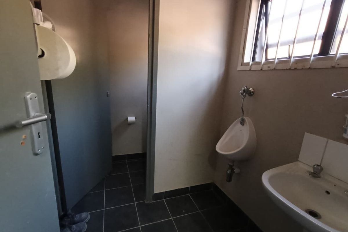 A functional industrial-style bathroom featuring a wall-mounted urinal, pedestal basin, and tiled flooring with security bars on the window.