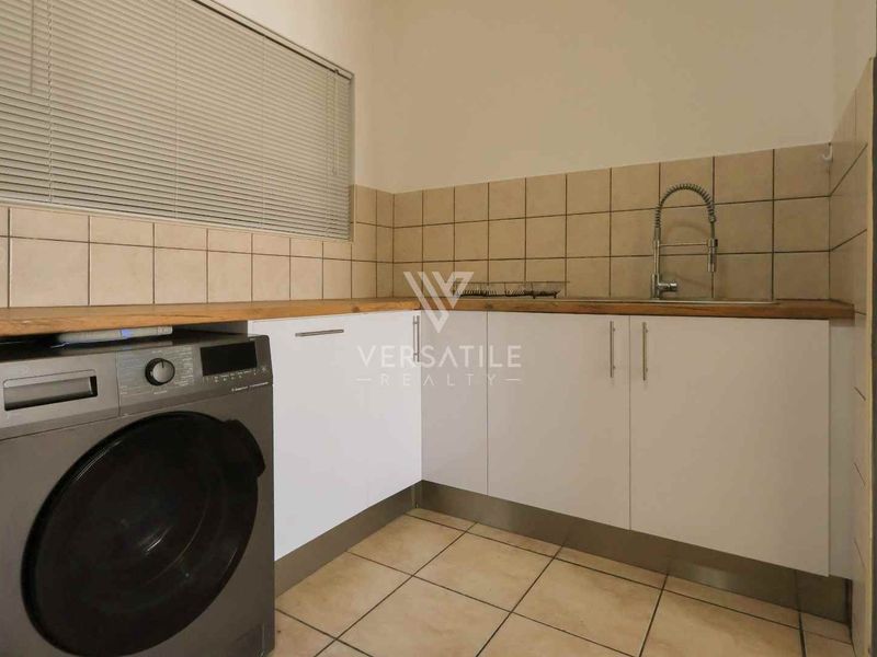 A functional laundry area featuring tiled backsplash, laminate countertops, base cabinetry, and a stainless steel sink with a window.