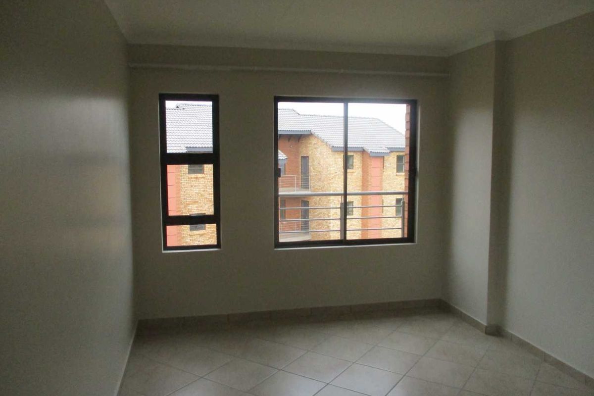 A bright, empty bedroom featuring neutral tiled flooring, large windows with metal frames, and a view of the surrounding apartment complex.