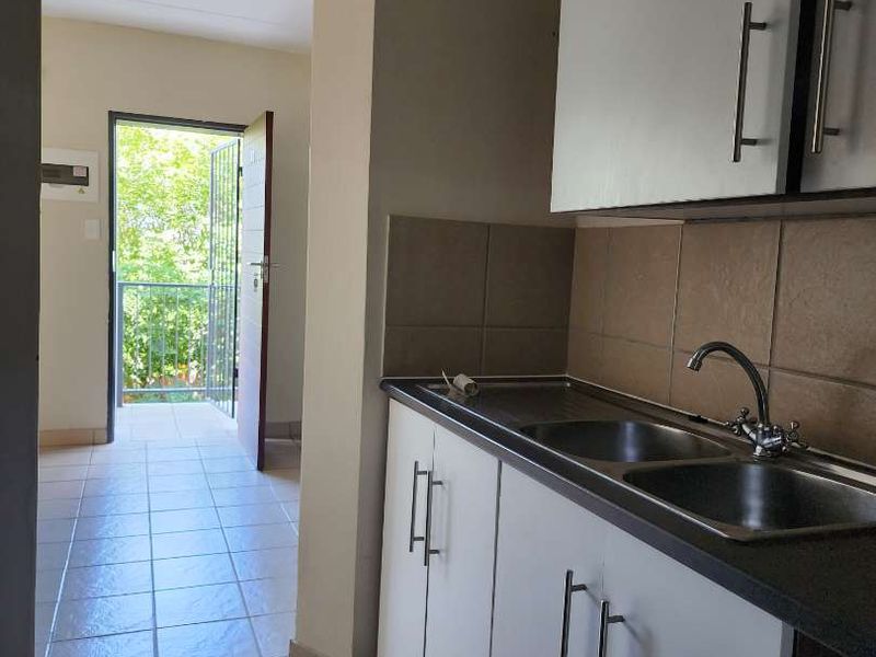 A compact kitchen area featuring white cabinetry, a stainless steel double sink, tiled splashback, and ceramic floor tiles, leading to an outdoor balcony.