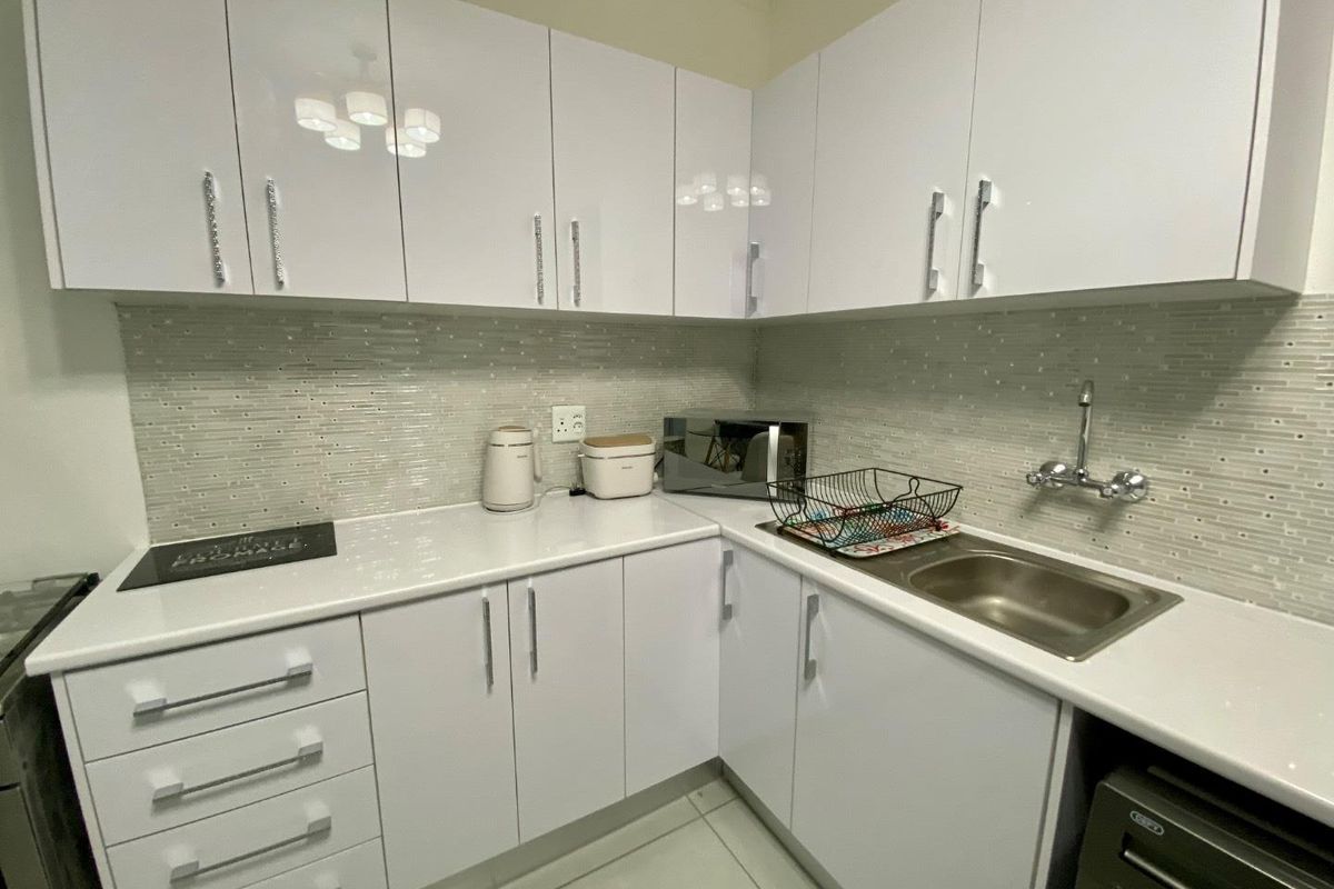 Modern kitchen featuring white high-gloss cabinetry, stone-look countertops, a mosaic tile backsplash, and a stainless steel sink with wall-mounted faucet.