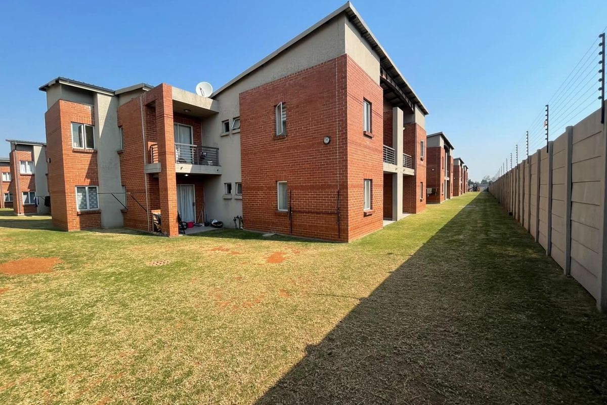 Rear view of a multi-story apartment complex featuring face brick construction, private balconies, and a large communal lawn enclosed by an electric-fenced boundary wall.