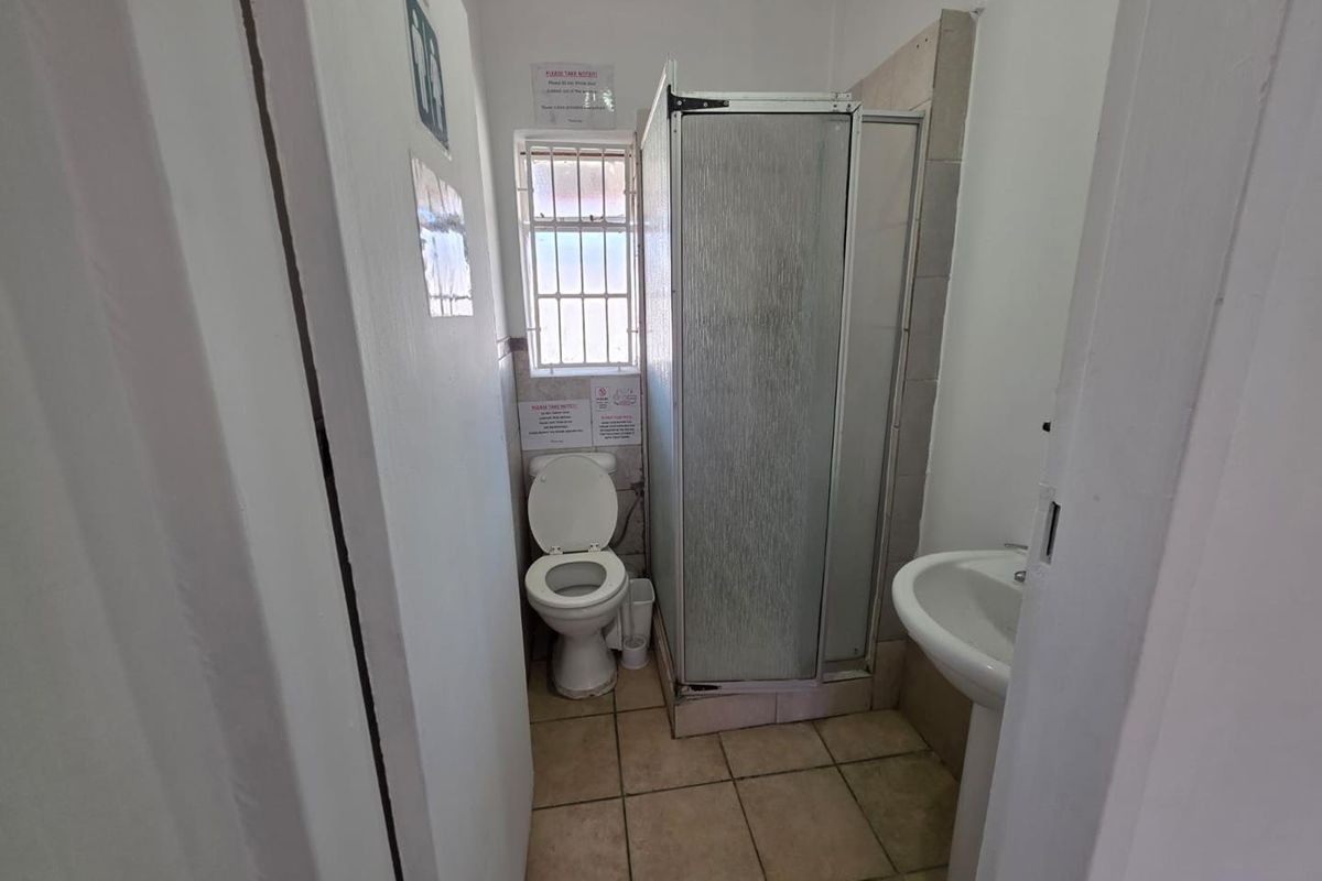 A compact bathroom featuring a shower cubicle, pedestal basin, and toilet, with tiled flooring and a window with security bars.