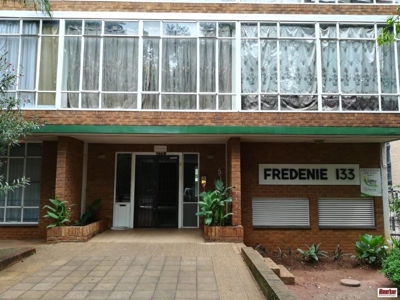 Front entrance of Fredenie 133 apartment building featuring a brick facade, large glass window panels, and a paved walkway with built-in planters.