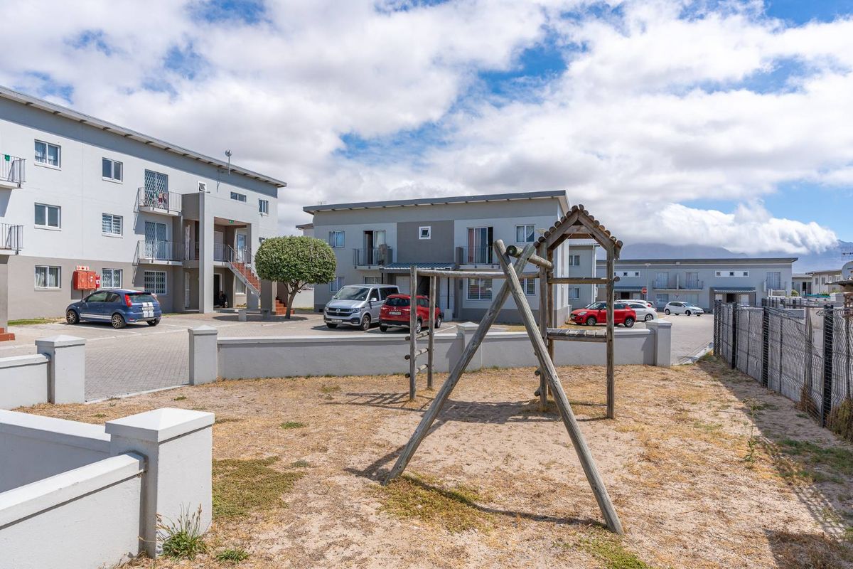 Exterior view of an apartment complex featuring a paved parking area, perimeter wall, and a communal playground space.