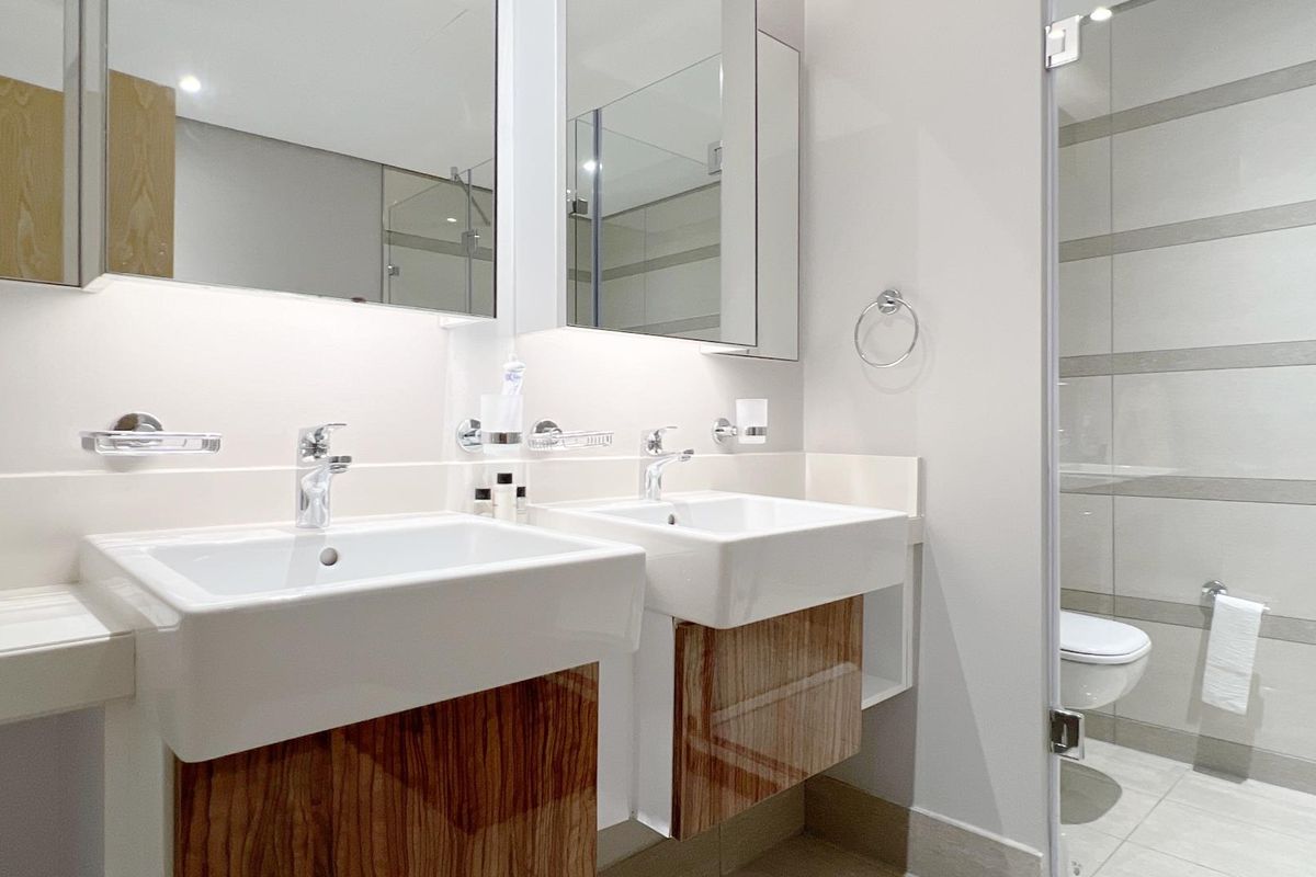 Modern bathroom featuring a double vanity with wood-finish cabinetry, integrated mirrors, and a separate glass-enclosed toilet area with neutral wall tiling.