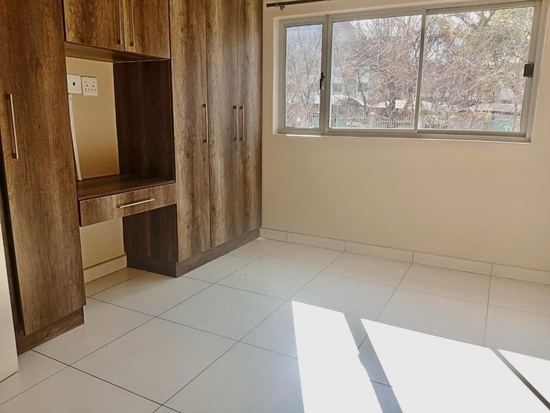 A bright, empty bedroom featuring modern wood-finish built-in cupboards, neutral tiled flooring, and large aluminium-framed windows allowing for ample natural light.