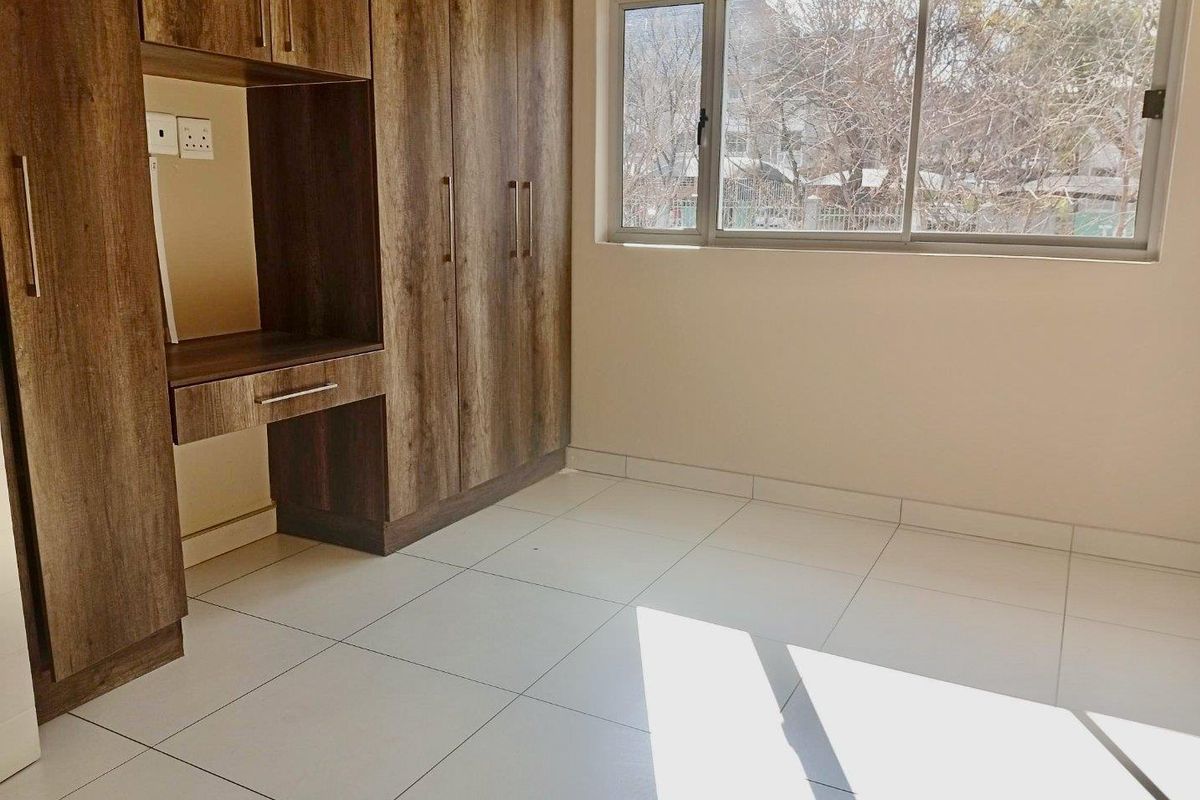 A bright, empty bedroom featuring modern wood-finish built-in cupboards, neutral tiled flooring, and large aluminium-framed windows allowing for ample natural light.