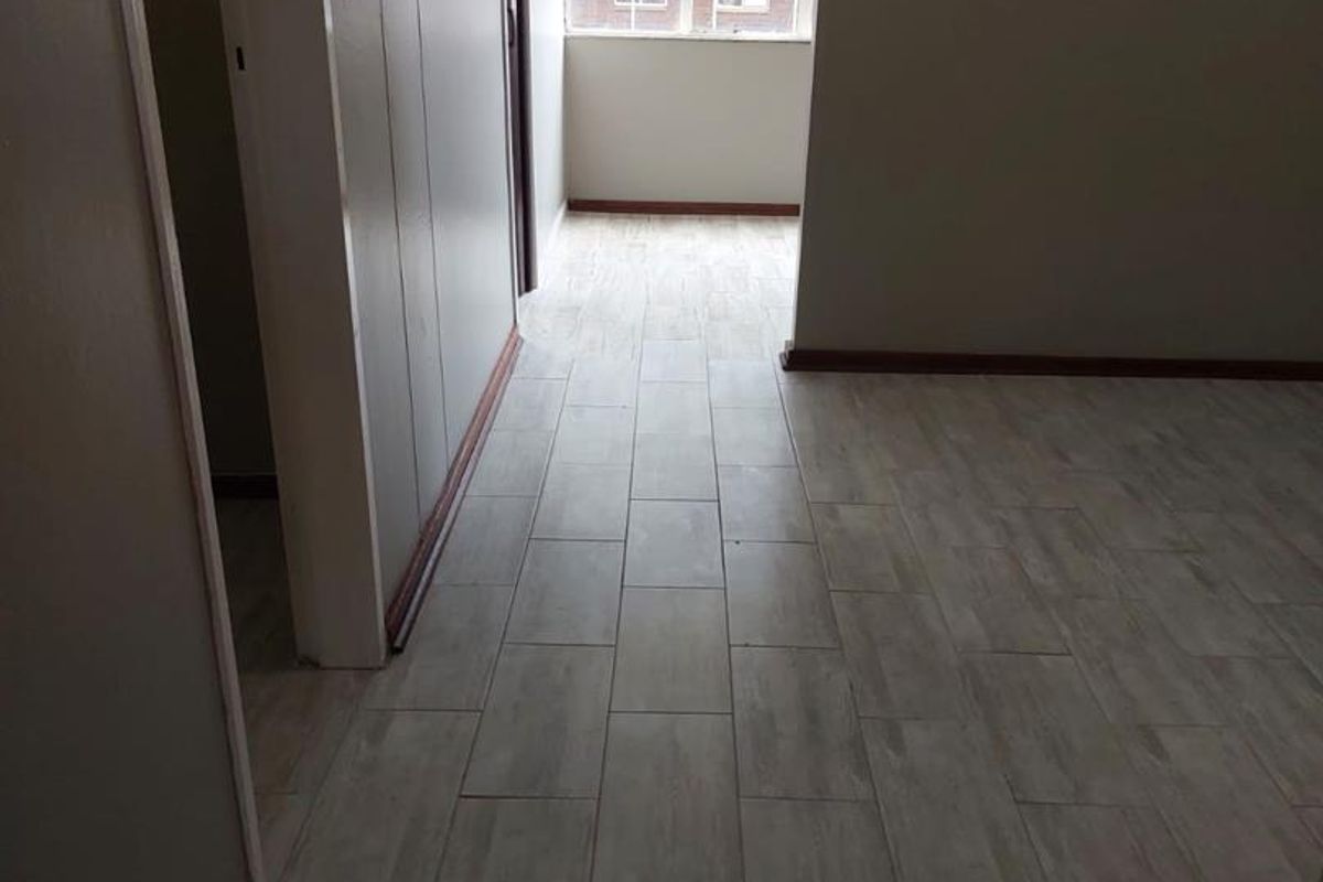 An empty interior space featuring light-toned wood-look floor tiles, neutral wall finishes, and a large window providing natural light.