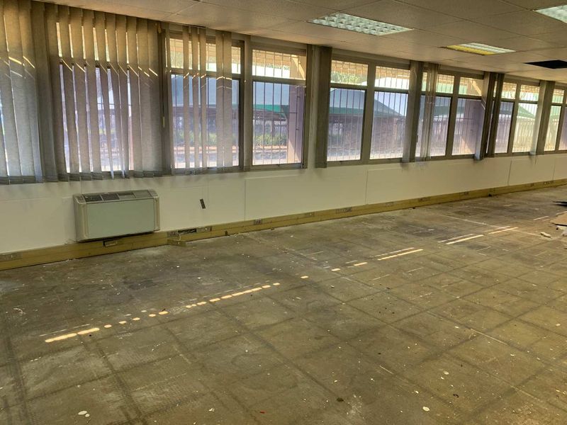 An empty industrial office space featuring large windows with security bars, wall-mounted air conditioning, and perimeter trunking, requiring refurbishment of flooring and ceiling.