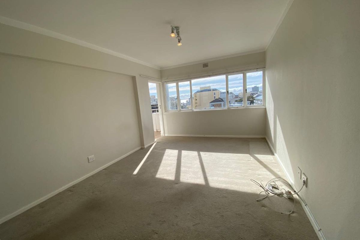 A bright, empty living room featuring large windows with urban views, neutral wall finishes, and carpeted flooring.