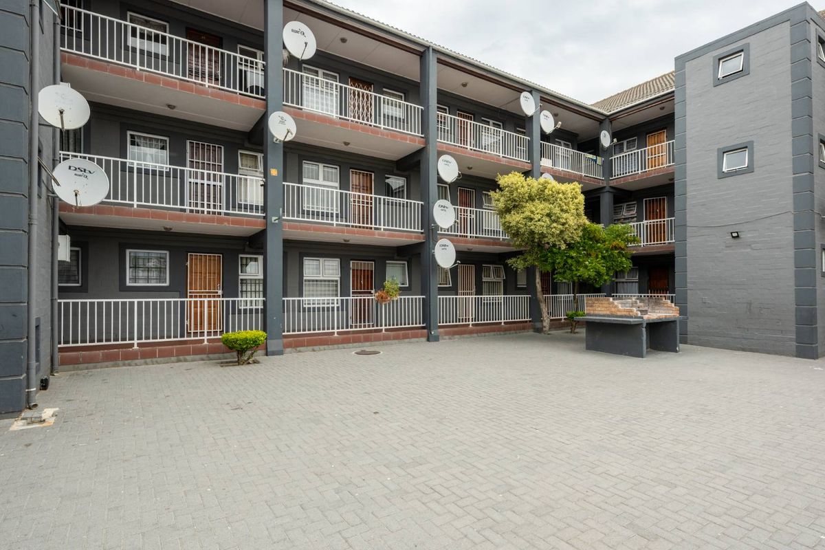 Exterior view of a multi-story apartment complex featuring paved courtyard, communal braai area, and balcony access for individual units.
