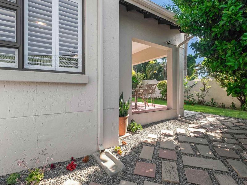 A covered patio area featuring paved flooring, adjacent to a well-maintained garden with a stone-paved walkway and exterior window shutters.