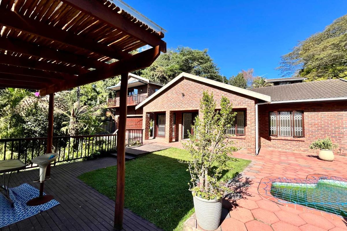 Rear exterior view featuring a brick house, timber pergola, wooden deck, paved patio area, and a swimming pool set within a garden.