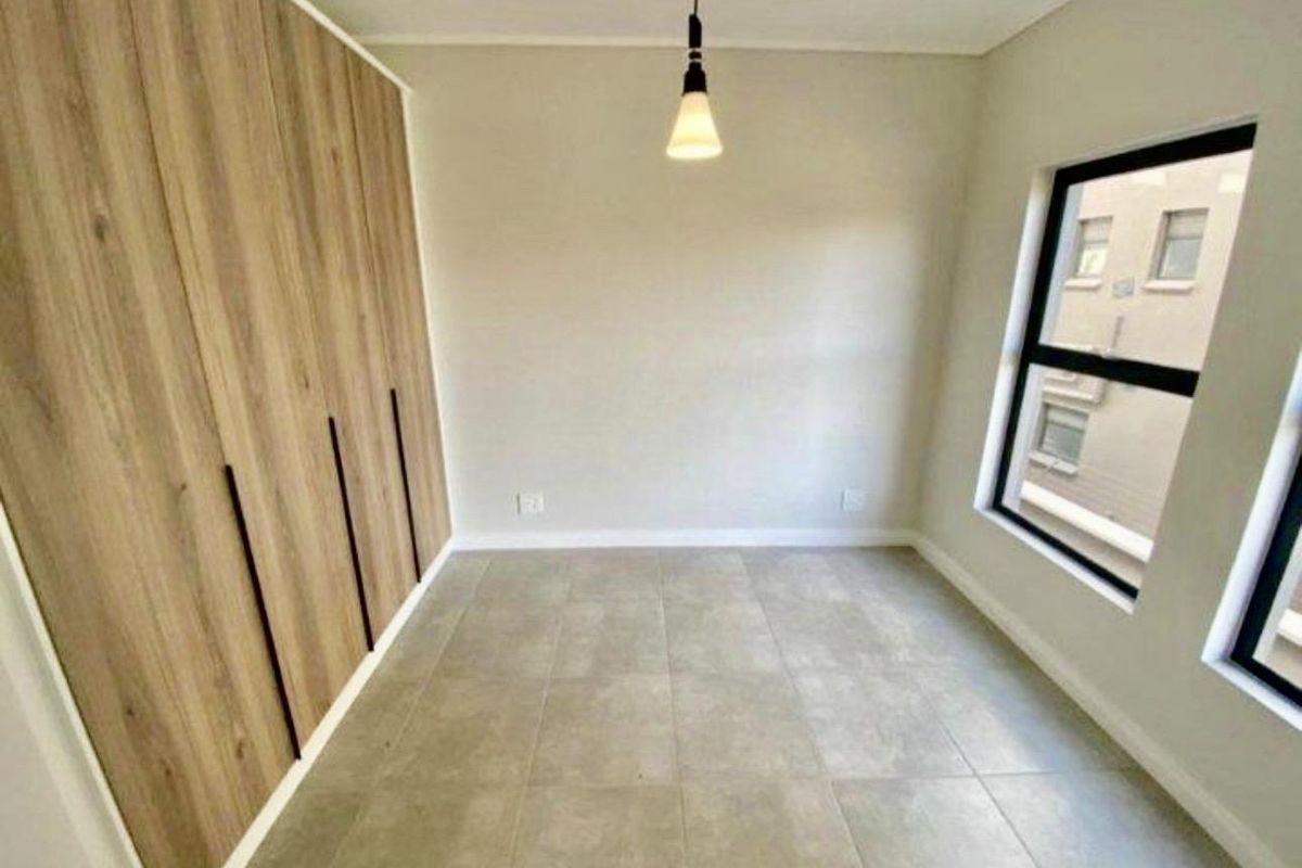 A bright bedroom featuring modern wood-finish built-in cupboards, neutral tiled flooring, and large windows providing natural light.