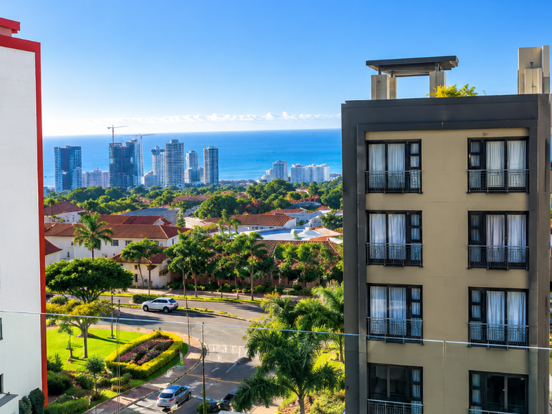 Elevated view overlooking the surrounding neighbourhood, landscaped gardens, and distant ocean horizon from a modern apartment balcony.