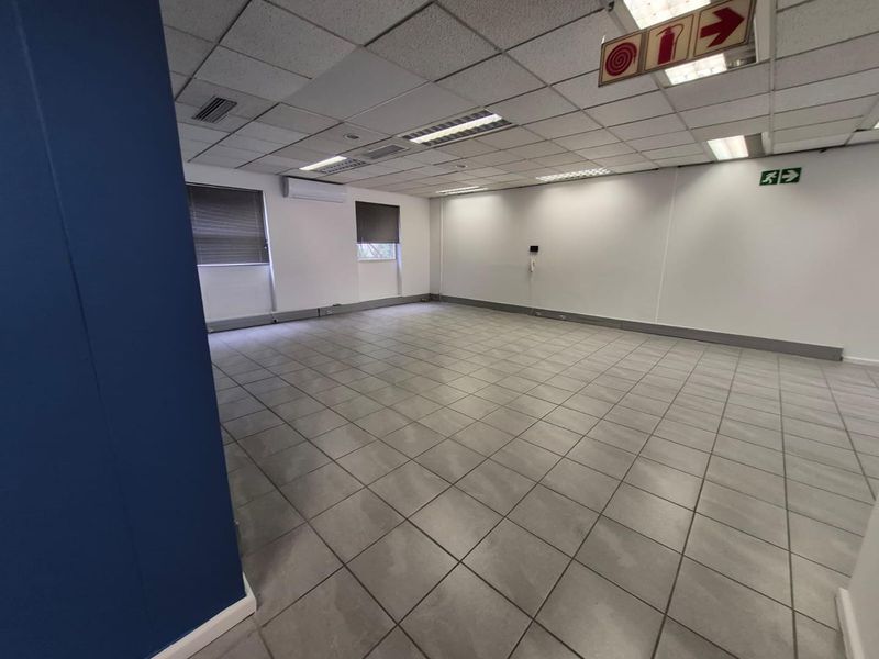 A spacious, empty office area featuring tiled flooring, a suspended ceiling with recessed lighting, and a wall-mounted air conditioning unit.