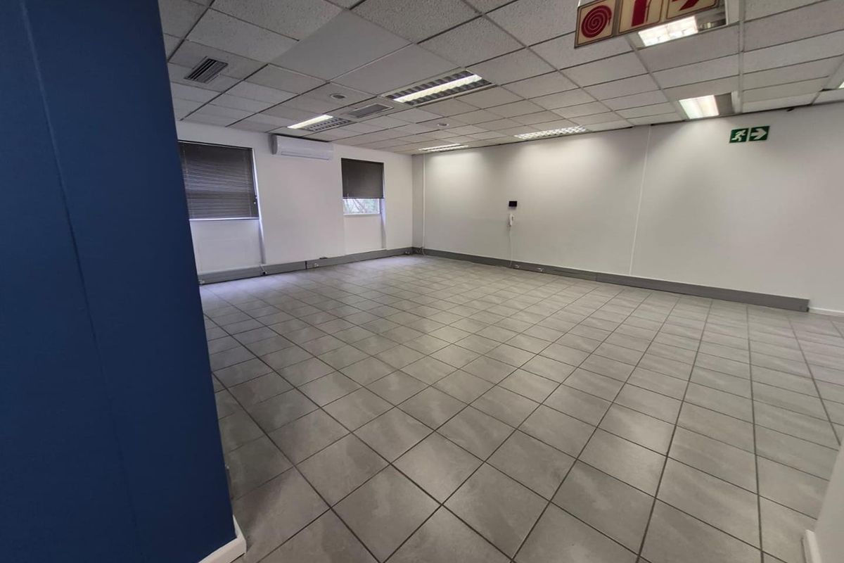 A spacious, empty office area featuring tiled flooring, a suspended ceiling with recessed lighting, and a wall-mounted air conditioning unit.