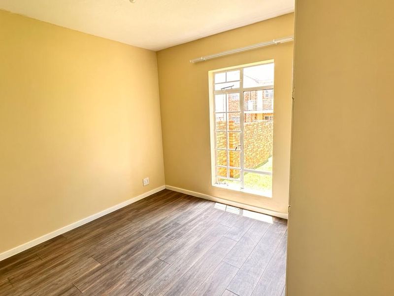 A bright, empty bedroom featuring modern laminate flooring, neutral wall paint, and a large window allowing for ample natural light.