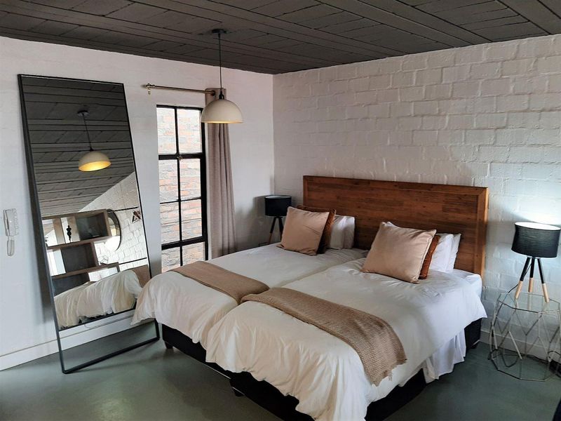 A well-maintained bedroom featuring a textured white brick wall, dark timber ceiling, and a large steel-framed window providing natural light.