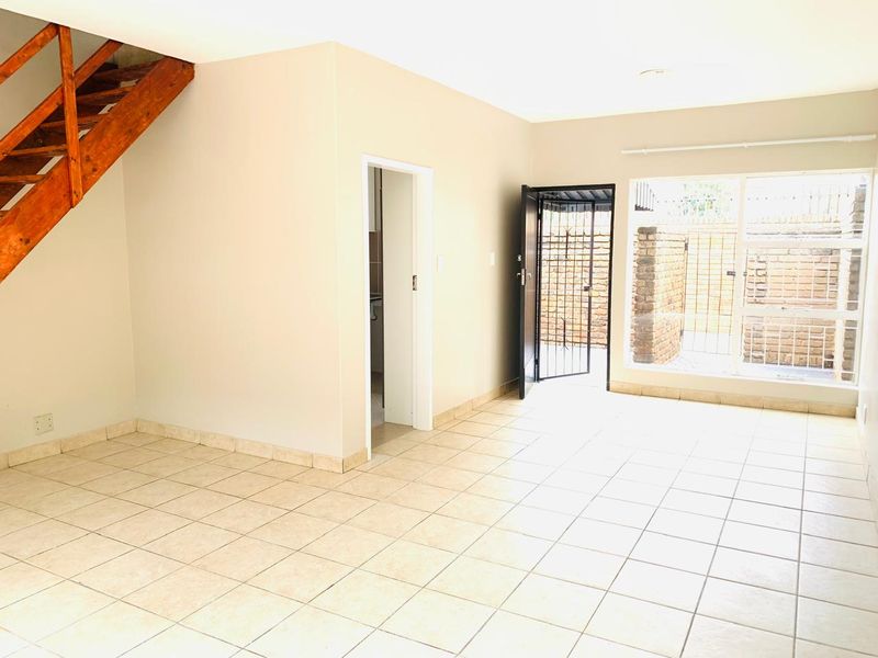 An open-plan living area featuring neutral tiled flooring, a wooden staircase, and direct access to an outdoor space through a security gate.
