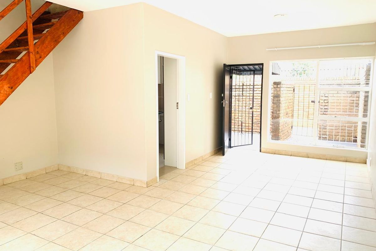 An open-plan living area featuring neutral tiled flooring, a wooden staircase, and direct access to an outdoor space through a security gate.