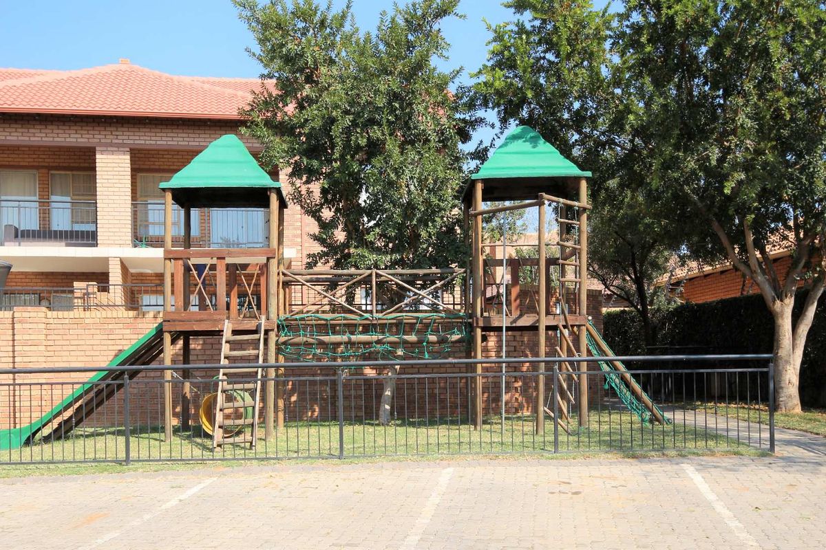 Exterior view of an apartment complex featuring a brick facade, balconies with metal railings, and a communal playground area with paved parking.