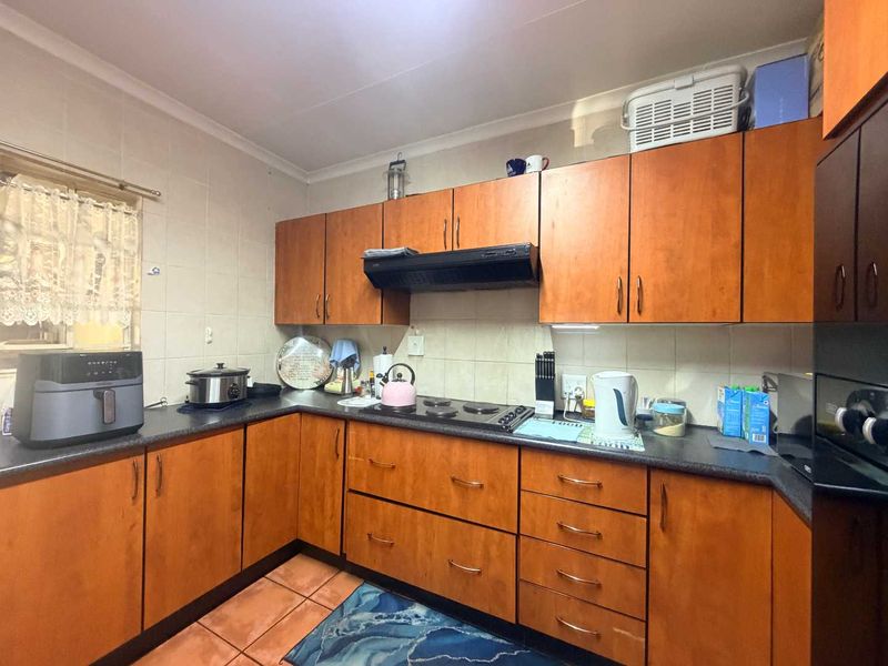 A functional kitchen featuring wooden cabinetry, dark laminate countertops, a built-in electric hob with extractor fan, and tiled finishes throughout.