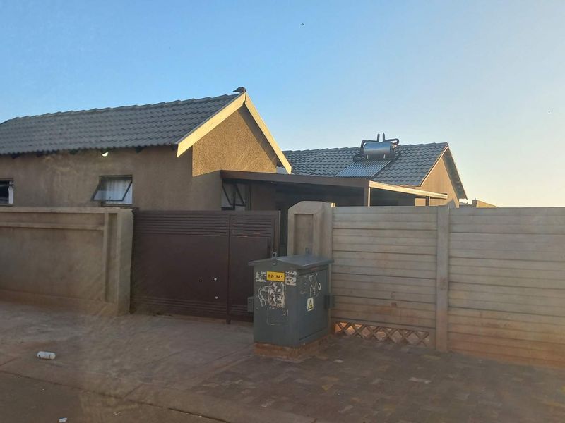 Front exterior view of a single-story house featuring a tiled roof with a solar geyser, enclosed by a precast concrete boundary wall and sliding gate.