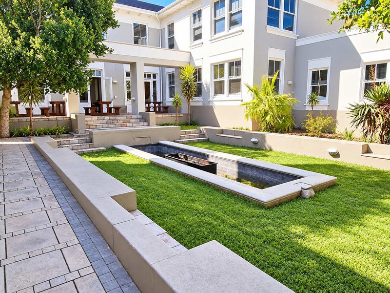 A well-maintained rear courtyard featuring a paved walkway, sunken water feature, tiered retaining walls, and manicured lawn area adjacent to the office building.