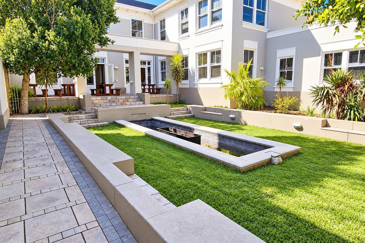 A well-maintained rear courtyard featuring a paved walkway, sunken water feature, tiered retaining walls, and manicured lawn area adjacent to the office building.