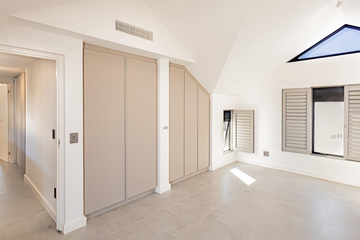 A bright, modern bedroom featuring built-in cupboards, tiled flooring, a skylight, and window shutters, with a view through to an adjacent hallway.