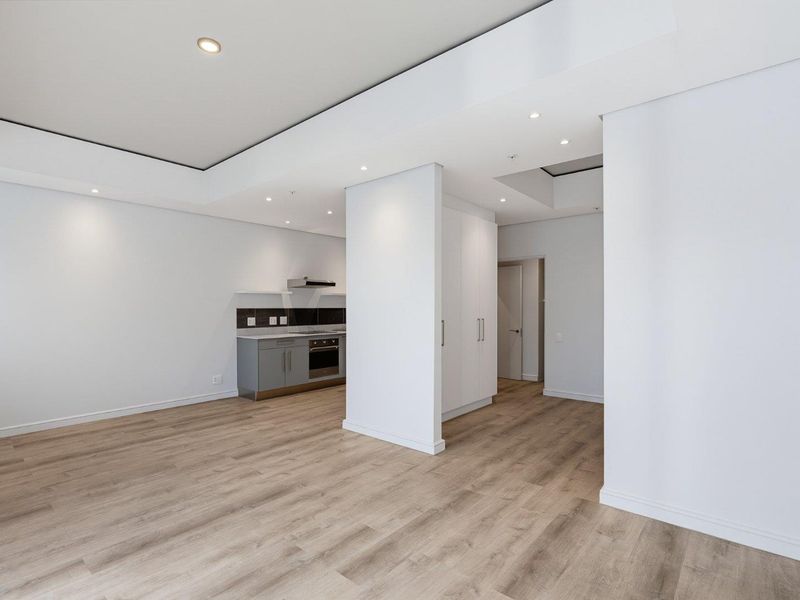 Modern open-plan living area featuring light laminate flooring, recessed ceiling lighting, and a compact kitchen with built-in cabinetry and integrated appliances.